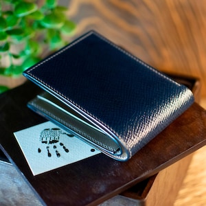 May include: A navy blue leather wallet with white stitching and a textured finish. The wallet is open, revealing a light-coloured interior. A small card with a handprint design rests beneath the wallet on a wooden surface.