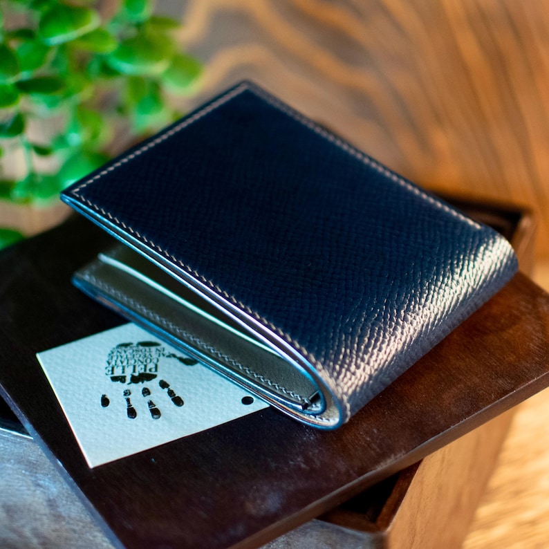 May include: A navy blue leather wallet with white stitching and a textured finish. The wallet is open, revealing a light-coloured interior. A small card with a handprint design rests beneath the wallet on a wooden surface.