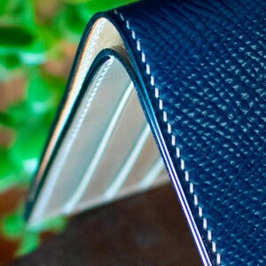 May include: A close-up shot of a navy blue leather wallet with white stitching. The wallet has a textured surface and a light-coloured interior. The edges are neatly finished, showcasing quality craftsmanship. The wallet is partially open, revealing the interior design.