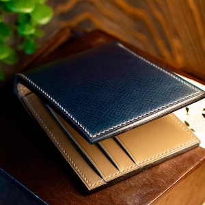 May include: A bi-fold wallet with a navy blue exterior and a tan interior. The wallet is made of textured leather and features white stitching around the edges. The interior has multiple card slots.