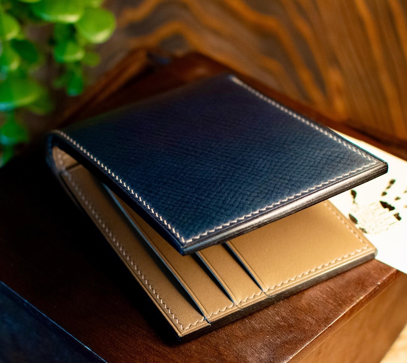 May include: A bi-fold wallet with a navy blue exterior and a tan interior. The wallet is made of textured leather and features white stitching around the edges. The interior has multiple card slots.