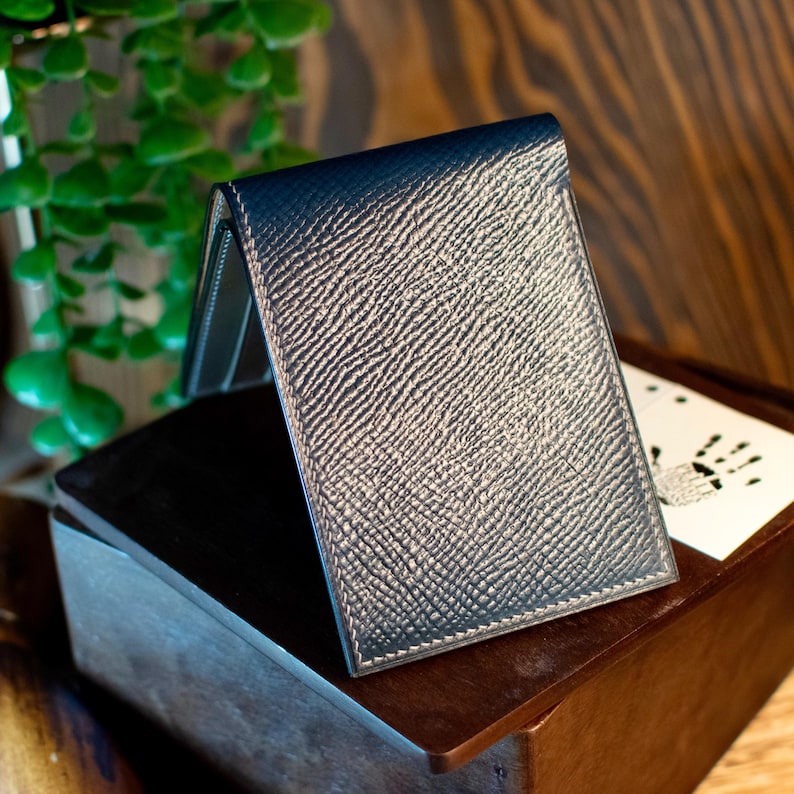 May include: A dark blue leather wallet with a textured finish, open to show a light grey interior. The wallet sits on a wooden box, with a small card visible in the background. The design is classic and sophisticated.