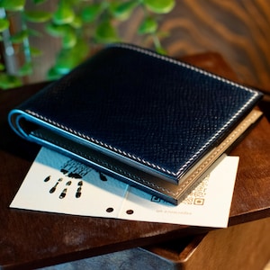 May include: A navy blue leather wallet with white stitching and a tan interior. The wallet is resting on a small white card with a QR code and a footprint design. The wallet is on a wooden surface.