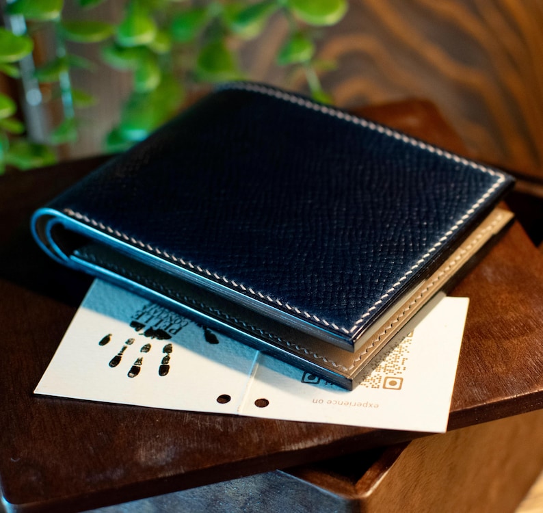 May include: A navy blue leather wallet with white stitching and a tan interior. The wallet is resting on a small white card with a QR code and a footprint design. The wallet is on a wooden surface.