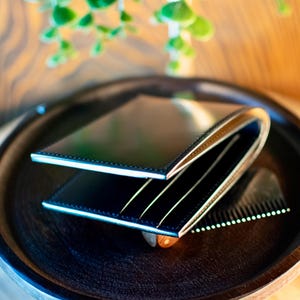 May include: A black leather wallet with a bi-fold design, featuring a contrasting light-colored interior. The wallet is open, revealing card slots. It rests on a dark wooden tray, alongside a comb. The overall aesthetic is minimalist and elegant.