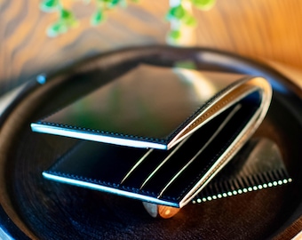 May include: A black leather wallet with a bi-fold design, showing a light-coloured interior. The wallet is open, revealing card slots. It sits on a dark wooden tray, with a comb. The overall aesthetic is minimalist and elegant.