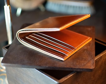 May include: A brown leather wallet with a bi-fold design. The wallet is open, showing several card slots and a contrasting interior. The edges are neatly stitched, and it rests on a dark wooden surface.