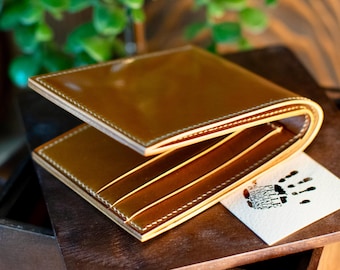 May include: A bi-fold leather wallet in a warm, golden-brown colour. The wallet has visible white stitching around the edges and multiple card slots. A small card with a handprint and text rests beside the open wallet.