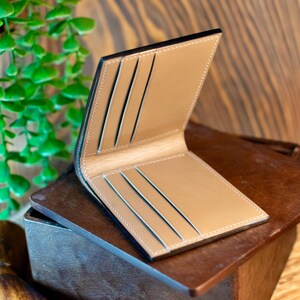 May include: Open, tan leather wallet with black edging and several card slots. The wallet is shown open, revealing its interior design and stitching. The background is a wooden surface.