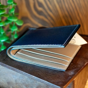 May include: A navy blue and tan leather bi-fold wallet. The wallet is open, showing card slots. White stitching details the edges. The wallet is displayed on a wooden surface. The wallet is a classic accessory.