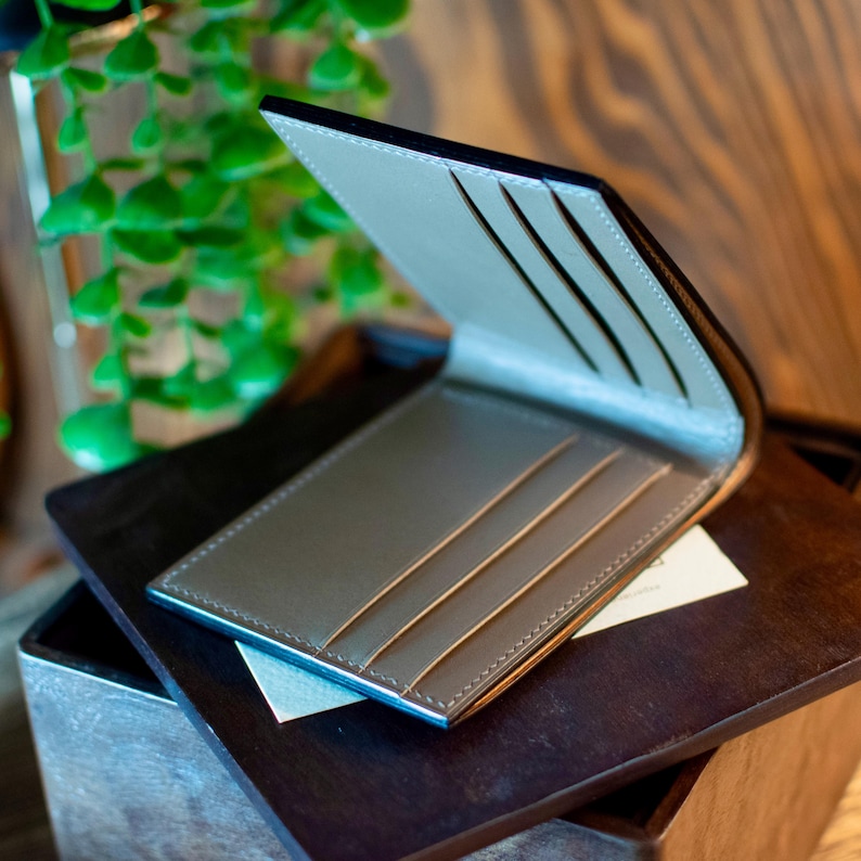May include: Open leather wallet with a light grey interior and card slots. The wallet has a black exterior and is displayed open, revealing the interior. The edges are stitched with a contrasting thread. The wallet is on a wooden surface.