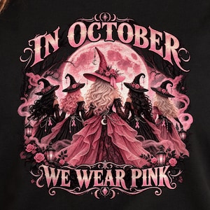 以下が含まれることがあります： 黒いTシャツに、魔女の帽子とフリルのドレスを身にまとった5人の人物が大きなピンクの月を背に立っているグラフィックが描かれています。テキストは「IN OCTOBER WE WEAR PINK」と書かれています。各ドレスにはピンクのリボンがあしらわれ、ランタンが配置されています。