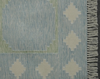 May include: A rectangular rug with a light blue and off-white geometric pattern. The rug features a diamond pattern and a fringe along one edge. The design incorporates light green accents, creating a textured, woven appearance. The rug is suitable for home decor.