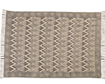 Puede incluir: Alfombra rectangular beige con un patrón de flechas negras repetidas. La alfombra tiene un borde con rayas verticales y flecos en los extremos cortos. Un revestimiento de suelo decorativo para un salón o dormitorio.