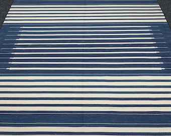 Peut inclure: Un tapis rectangulaire avec un motif répétitif de rayures horizontales bleu marine et blanc cassé. Les rayures sont espacées uniformément, avec un élément de design unique au centre où certaines rayures semblent s'estomper. Le tapis a une bordure frangée.
