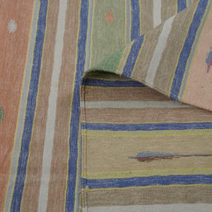 Puede incluir: Una alfombra a rayas multicolor con una esquina doblada. La alfombra presenta rayas verticales en tonos melocotón, azul, marrón, blanco y verde. La esquina doblada revela la parte inferior de la alfombra, mostrando un patrón de rayas similar.