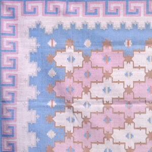 May include: Close-up of a patterned textile. The design includes a pink and light blue Greek key border. The central pattern features white, pink, and blue geometric shapes with star and cross-like details. The colour scheme is soft and pastel, creating a delicate aesthetic.