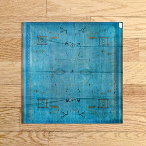 Op de afbeelding: Een blauw tapijt met een geometrisch patroon in tinten bruin en zwart. Het tapijt heeft een gefranst randje en is dubbelgevouwen.