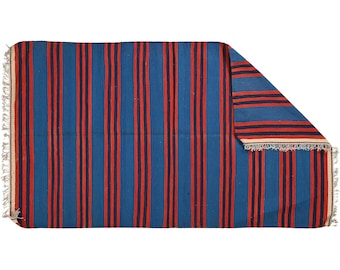 May include: A rectangular rug with a blue background and vertical stripes in red and navy. The rug has a fringed edge on one side. The corner of the rug is folded over, revealing the striped pattern on the underside.