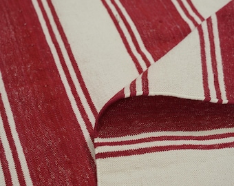 Puede incluir: Una alfombra a rayas rojas y blancas con una esquina doblada. La alfombra presenta gruesas rayas rojas verticales que se alternan con rayas blancas más anchas. La tela parece tejida, con una superficie ligeramente texturizada. La esquina doblada revela la parte inferior de la alfombra.