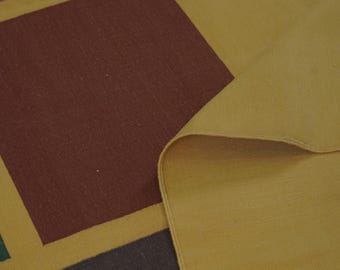 May include: A close-up of a woven rug with a geometric pattern. The rug features large squares in shades of brown, green, and red, set against a mustard yellow background. The corner of the rug is folded over, revealing the underside.