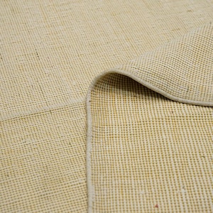 May include: Close-up of a cream-colored woven rug with a subtle texture. The rug has a white border and is folded over on itself.