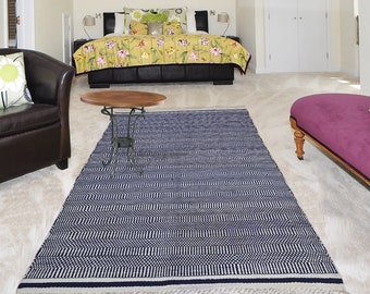 Puede incluir: Una alfombra azul y blanca con estampado y flecos en los bordes. La alfombra está colocada sobre una alfombra beige claro en una habitación con una cama, un sofá y una mesa auxiliar.