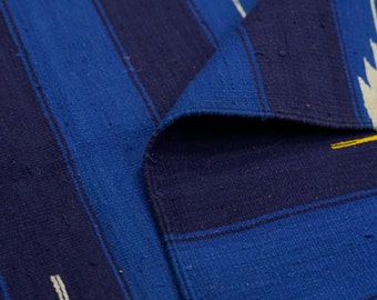 Puede incluir: Un tejido con rayas verticales en varios tonos de azul oscuro y azul real. La tela presenta un tejido texturizado visible con finos acentos lineales en color blanco roto y amarillo que aparecen como trazos cortos e irregulares en la superficie.
