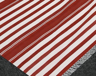 Puede incluir: Un textil a rayas rojas y blancas con un borde con flecos. Las rayas diagonales alternan bandas anchas rojas y blancas. El textil es probablemente una alfombra o una manta.