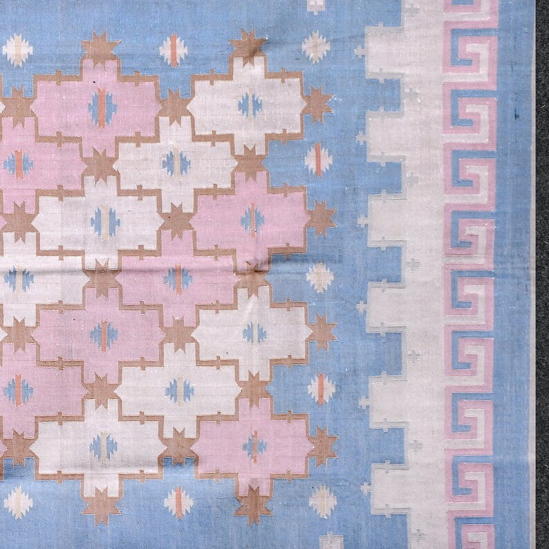 May include: A close-up of a woven textile with a geometric pattern. The design features a repeating pattern of pink, white, and tan shapes on a light blue background. The border has a Greek key design in pink and white.