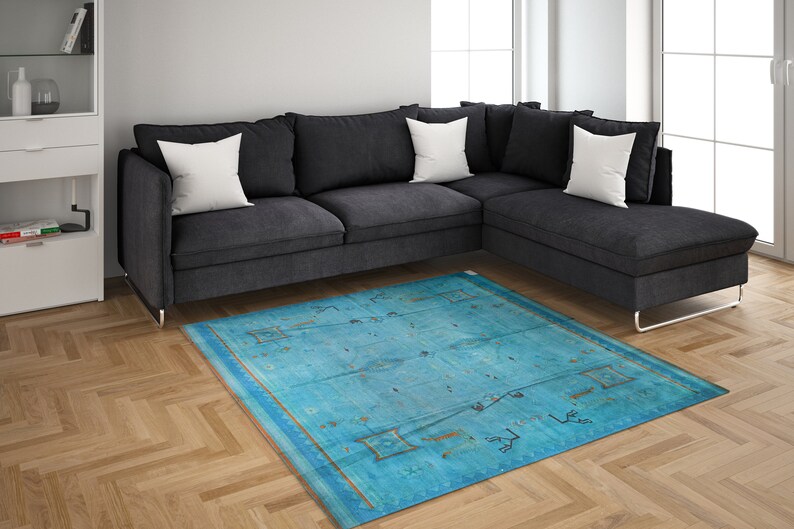 Op de afbeelding: Een blauw en gouden tapijt met een geometrisch patroon. Het tapijt ligt voor een grijze hoekbank met witte kussens.