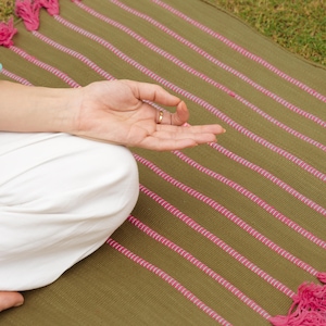 Puede incluir: Una esterilla de yoga verde oliva con rayas rosas y borlas. Una persona está sentada en la esterilla en posición meditativa, con la mano en un mudra. La esterilla está diseñada para yoga y meditación.