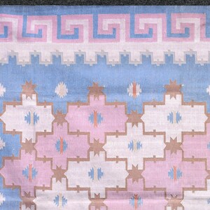 May include: Close-up of a textile with a repeating geometric pattern. The design features a light blue background with pink, white, and tan shapes. A Greek key border in pink and white accents the top edge. The overall aesthetic is reminiscent of a rug or fabric.