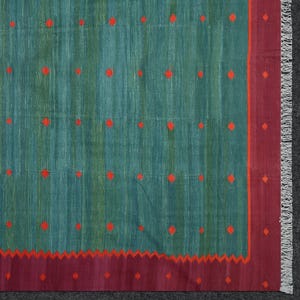 Op de afbeelding: Een turquoise en groen tapijt met een herhalend patroon van rode ruitvormen. Het tapijt heeft een bordeauxrode rand met een rood zigzagontwerp. De rand van het tapijt heeft franjes.