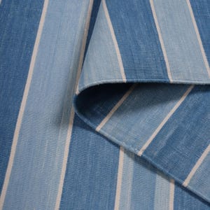 May include: A blue and white striped fabric, with varying shades of blue and white stripes. The fabric is folded over, revealing the underside. The stripes are vertical and evenly spaced, creating a clean and classic design. The fabric appears to be a woven material.