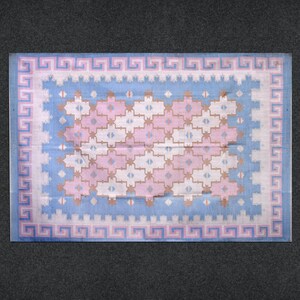 May include: A rectangular rug with a light blue and pink geometric pattern. The rug features a border with a Greek key design in pink and white. The central design includes pink, white, and tan cross-like shapes.