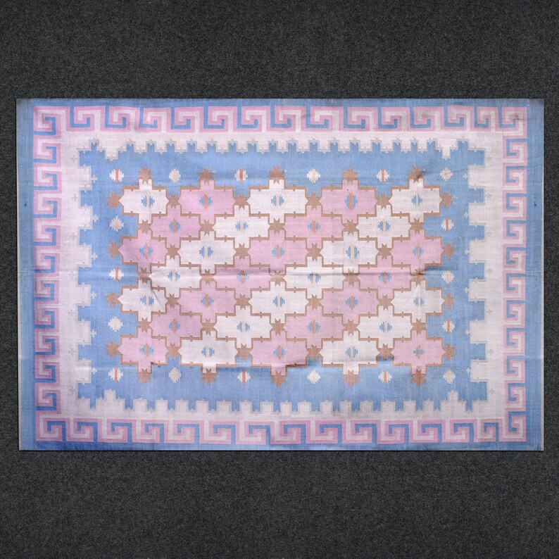 May include: A rectangular rug with a light blue and pink geometric pattern. The rug features a border with a Greek key design in pink and white. The central design includes pink, white, and tan cross-like shapes.