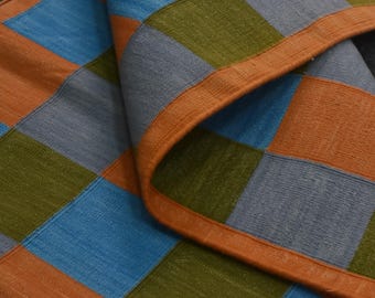 May include: A woven textile featuring a repeating checkered pattern in shades of orange, blue, olive green, and gray. The fabric has a visible textured weave and is finished with a solid orange border along the folded edge of the material.