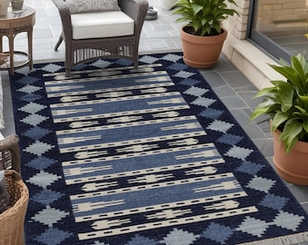 May include: A rectangular outdoor rug with a geometric pattern in shades of navy blue, light blue, and cream. The rug features a diamond border and horizontal stripes, with a fringed edge. The rug is placed on a patio with a wicker chair and potted plants.