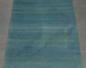 May include: A rectangular area rug featuring a solid teal and blue gradient color scheme with subtle horizontal bands. The rug has a flat-woven texture and is finished with short, white fringe detailing along the top and bottom edges.
