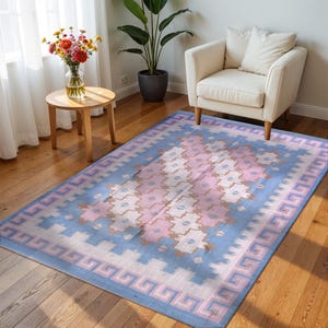 May include: A rectangular rug with a light blue backdrop and a central pattern of pink and white geometric shapes. The rug features a border with a Greek key design in pink and blue. It's placed on a wooden floor.