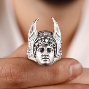 May include: A silver ring with a detailed design of a winged figure wearing a helmet. The figure has a serious expression and is looking straight ahead.