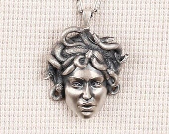 May include: A silver-toned pendant necklace featuring a detailed depiction of Medusa's face. The pendant showcases Medusa with snakes for hair, attached to a silver chain. The necklace is displayed against a textured, off-white background.