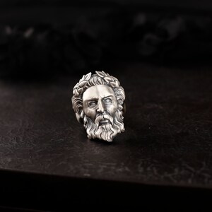 May include: A silver ring with a detailed design of a man's face with a beard and a laurel wreath on his head.