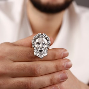 May include: A silver ring with a detailed face sculpture, featuring a beard and a crown of leaves.