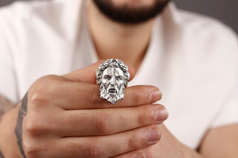 May include: A silver ring with a detailed face sculpture, featuring a beard and a crown of leaves.