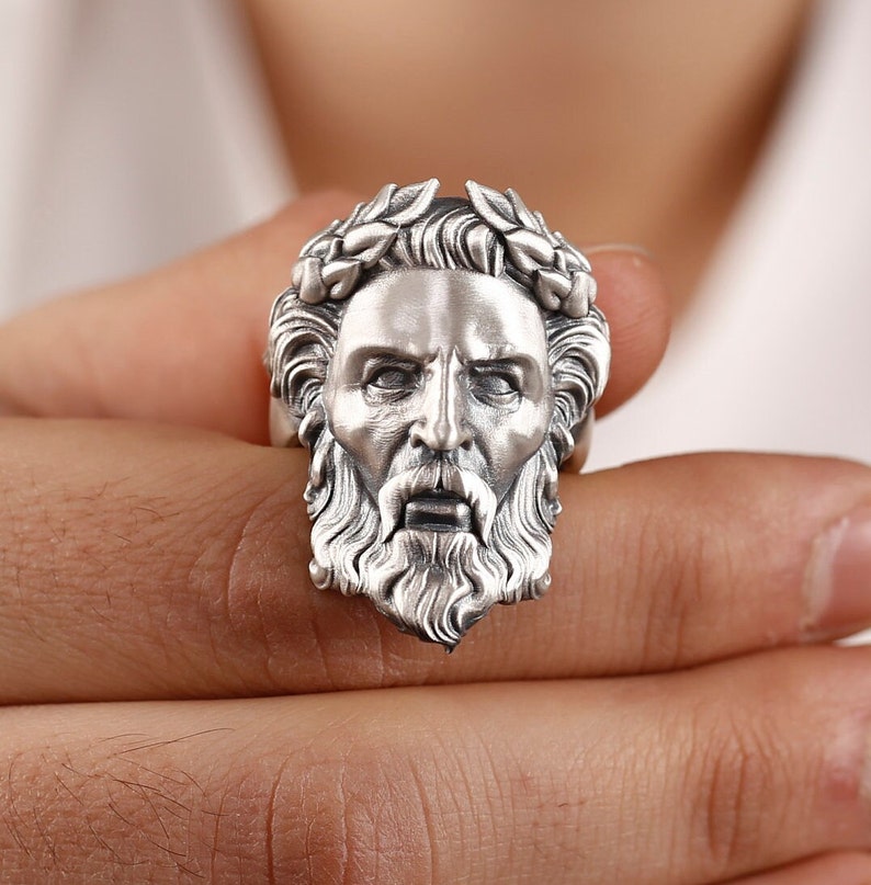 May include: A silver ring with a detailed sculpture of a man's face with a laurel wreath. The man has a beard and mustache.