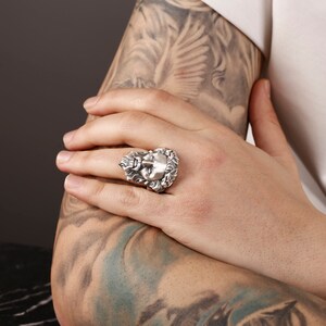 May include: A silver ring with a detailed design of a face surrounded by leaves.
