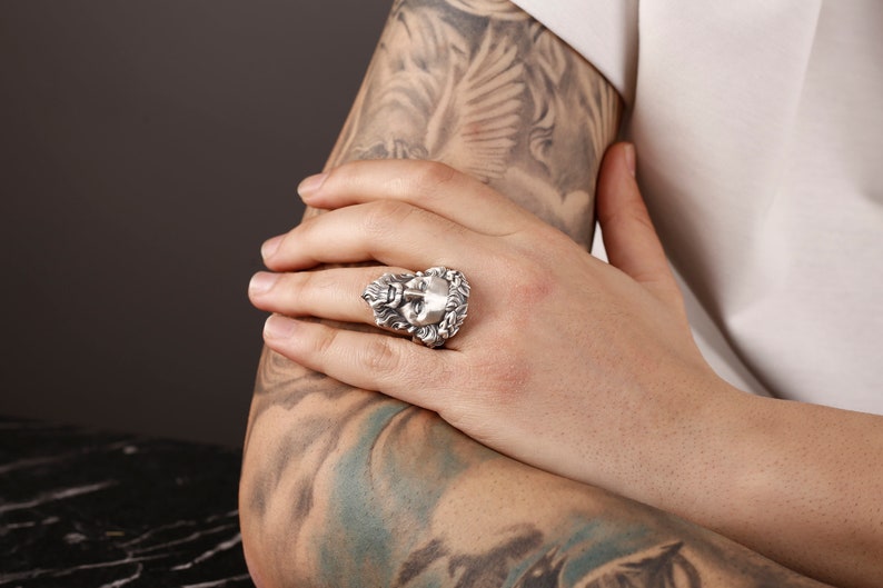 May include: A silver ring with a detailed design of a face surrounded by leaves.
