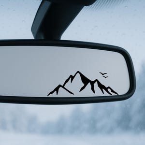 Könnte beinhalten: Schwarze Bergkette und zwei Vögel Silhouette Aufkleber auf dem Rückspiegel eines Autos. Das minimalistische Design ist ideal für Outdoor-Enthusiasten. Das Bild ist vor einem verschwommenen, neutralen Hintergrund.
